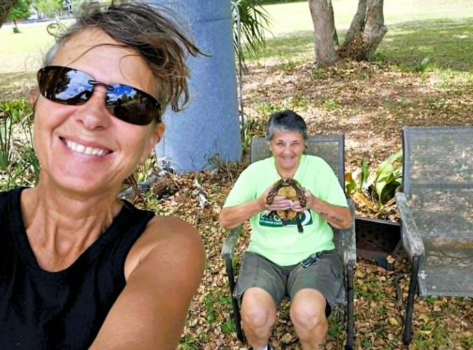 Two women smiling for a selfie outdoors, one holding a turtle. They are surrounded by greenery and seated on chairs.